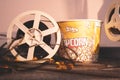 Old-school projector reel with uncoiled film is leaning against paper bucket of caramel popcorn in side warm light of