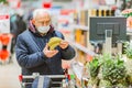 Old senior european man wearing protective facial mask looking at banana in the supermarket. Shopping during COVID-19 concept