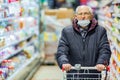 Old senior european man wearing protective facial mask pushing shopping cart in the supermarket. Shopping during COVID-19 concept