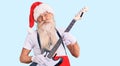 Old senior man with grey hair and long beard wearing santa claus costume playing electric guitar looking positive and happy Royalty Free Stock Photo