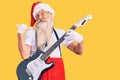 Old senior man with grey hair and long beard wearing santa claus costume playing electric guitar pointing thumb up to the side Royalty Free Stock Photo