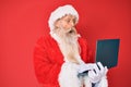Old senior man with grey hair and long beard wearing santa claus costume using laptop afraid and shocked with surprise and amazed Royalty Free Stock Photo