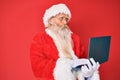 Old senior man with grey hair and long beard wearing santa claus costume using laptop making fish face with mouth and squinting Royalty Free Stock Photo