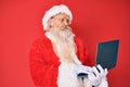 Old senior man with grey hair and long beard wearing santa claus costume using laptop relaxed with serious expression on face Royalty Free Stock Photo