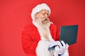 Old senior man with grey hair and long beard wearing santa claus costume using laptop sticking tongue out happy with funny Royalty Free Stock Photo