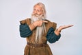 Old senior man with grey hair and long beard wearing viking traditional costume amazed and smiling to the camera while presenting