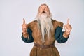 Old senior man with grey hair and long beard wearing viking traditional costume amazed and surprised looking up and pointing with