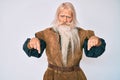 Old senior man with grey hair and long beard wearing viking traditional costume pointing down looking sad and upset, indicating