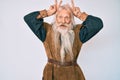 Old senior man with grey hair and long beard wearing viking traditional costume posing funny and crazy with fingers on head as