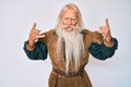 Old senior man with grey hair and long beard wearing viking traditional costume shouting with crazy expression doing rock symbol