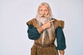 Old senior man with grey hair and long beard wearing viking traditional costume surprised pointing with finger to the side, open