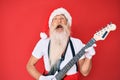 Old senior man wearing santa claus costume playing electric guitar angry and mad screaming frustrated and furious, shouting with Royalty Free Stock Photo