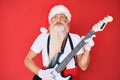 Old senior man wearing santa claus costume playing electric guitar puffing cheeks with funny face Royalty Free Stock Photo
