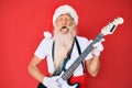 Old senior man wearing santa claus costume playing electric guitar sticking tongue out happy with funny expression Royalty Free Stock Photo