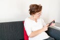 Old senior woman female sit home flat couch hold old mobile phone write text watch video call alone unhappy sad thinking