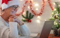 Old senior woman laughing using laptop computer in video call wearing santa hat. Christmas decorations at home with lights. New Royalty Free Stock Photo