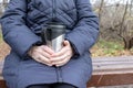 An old senior woman sitting on a bench in late autumn park and holding thermo cup in hands, hot drinks outdoor concept Royalty Free Stock Photo