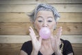 Old senior young woman do pink bubble gum ball surpsrised. Portrait of mature trendy female people with wooden background. Happy