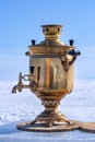 An old shiny copper and brass samovar stands on a cut of a tree trunk on an ice plate on a background of snow. Royalty Free Stock Photo