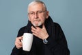 Old sick man with gray hair and beard in eyeglasses and scarf with big cup with tea holding hand near throat