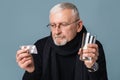 Old sick man with gray hair and beard in eyeglasses and scarf thoughtfully looking on pills holding glass of water in Royalty Free Stock Photo