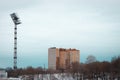 An old stadium lamp high in the cloudless sky. Tall residential building in the distance and lots of leaveless trees in the bottom