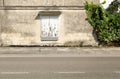 Old stained plaster wall with a peeled window shutter at the roadside. Concrete sidewalk and asphalt road in front, ivy by side. Royalty Free Stock Photo