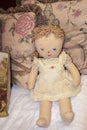 Old stained rag doll with painted eyes and mouth and embroidered searsucker pinafore propped against floral fringed pillows -