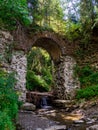 Old stone arched destroyed bridge over small river in a mountain forest