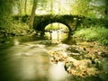 Old stony bridge above autumn river. Water of stream full of colorful leaves, leaves on gravel, blue blurred water is running over Royalty Free Stock Photo