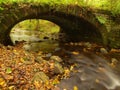 The old stony bridge above stream. Water of brook full of colorful leaves, leaves on gravel, blue blurred water is running.