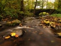 The old stony bridge above stream. Water of brook full of colorful leaves, leaves on gravel, blue blurred water is running.