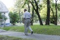 An old, stooped man walks sullenly and alone along the path in the park with a gray umbrella instead of a cane.