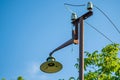 An old street lamp on a rusty pole with a broken bulb and torn wires against a background of green leaves and blue sky. Royalty Free Stock Photo