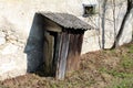 Old traditional outdoor wooden toilet with cracked dilapidated wooden boards and broken doors attached to concrete and stone wall