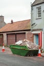 Old weathered green skip full with household rubbish on a street and safety red cones in old town. House renovation concept.