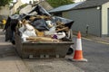 Old weathered green skip full with household rubbish on a street and safety red cones in old town. House renovation concept.