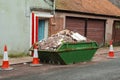 Old weathered green skip full with household rubbish on a street and safety red cones in old town. House renovation concept.