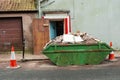 Old weathered green skip full with household rubbish on a street and safety red cones in old town. House renovation concept.