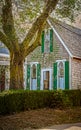 Old weathered vintage wooden shingled house with arched church-like windows and green shutters stand behind tree and hedge Royalty Free Stock Photo