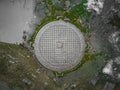 An old wet metal manhole on an abandoned asphalt road overgrown with moss and puddles and remnants of wet snow around. Top view.