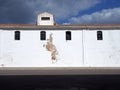 Old white painted building with high small windows and a tiled roof alongside a black roof with bright sunlit cloudy blue sky Royalty Free Stock Photo