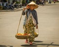 Old woman carries a heavy load of fruit in baskets for sale.