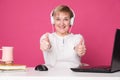 Old woman in her 60s works on computer, wearing headphohes. Laptop on white table and Pink background. She make thumbs up gesture