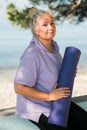 Old woman holding yoga mat and preparing to practice yoga or meditation outdoors on sea beach. Happy mature woman with Royalty Free Stock Photo