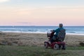 An old woman rides on a electric powered wheelchair parked on the beach at sunset time, in a lonely atmosphere. Lonely widow old