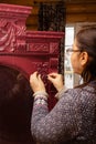 Old woman sticking carved from wood ornaments to wooden red cardboard color with round glass in front in wooden cottage