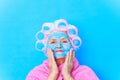 old woman wirh pink curlers on head and blue hyaluronic beauty mask on face wear cotton robem preparing for a date