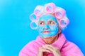 old woman wirh pink curlers on head and blue hyaluronic beauty mask on face wear cotton robe, preparing for a date