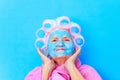 old woman wirh pink curlers on head and blue hyaluronic beauty mask on face wear cotton, robe preparing for a date